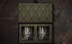 Two glass tumblers in a decorative black and gold box on a dark wooden surface.