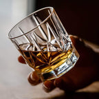 Hand holding a crystal glass filled with whiskey against a neutral background