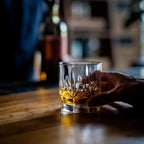 Hand holding a glass of whiskey on a wooden surface with blurred background