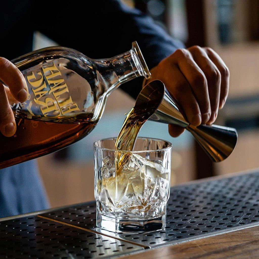Person pouring a clear liquid from a bottle into a glass with ice on a bar counter.